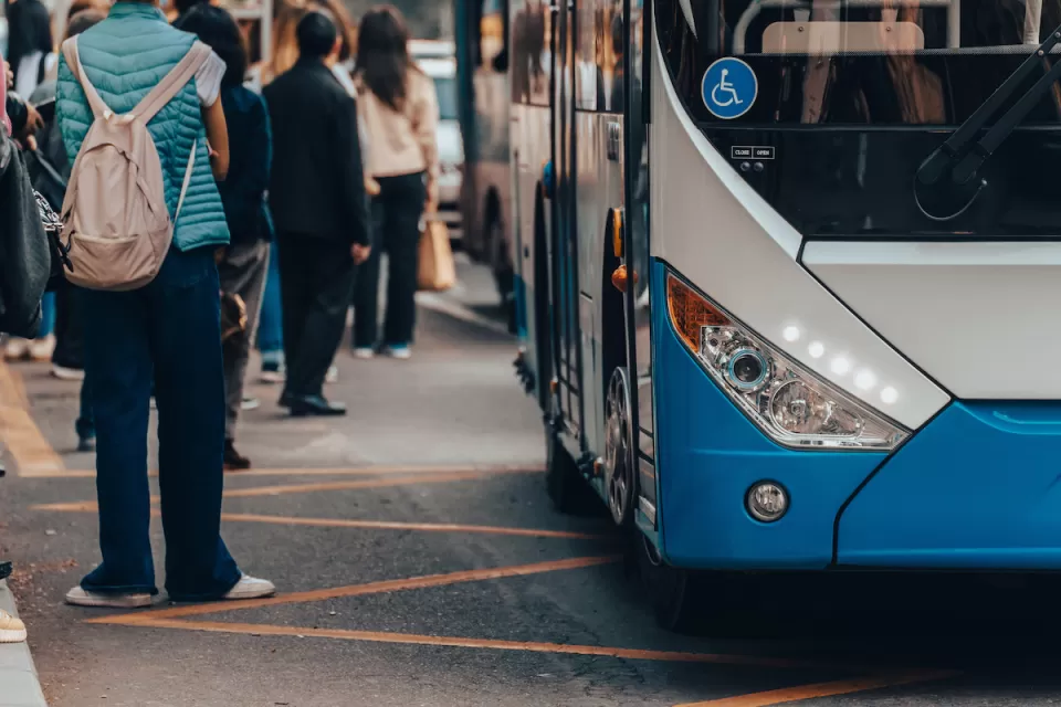 Close-up of front side of blue and white bus with accessibility sticker and people wearing backpacks waiting outside of bus.